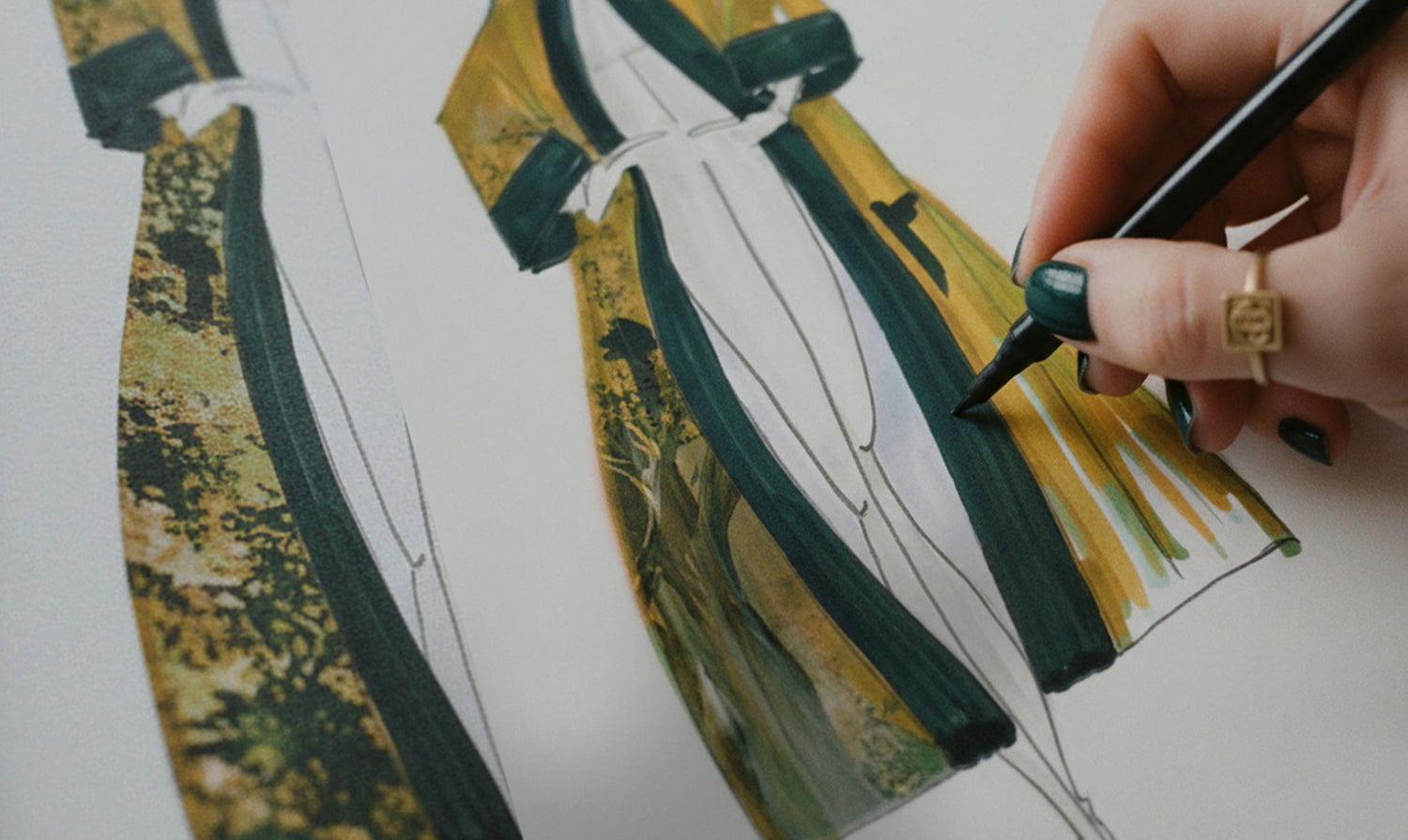 Designer's hand with dark teal nail polish and gold signet ring drawing kimono fashion illustration with brush pen