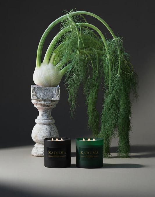 KARUMA black and green luxury scented candles with gold lettering displayed in front of wild fennel plant and cracked stone pedestal, editorial still life product photography, Black Forest luxury home fragrance