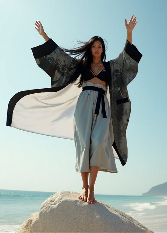Woman in traditional outfit standing on a rock by the ocean with arms raised.