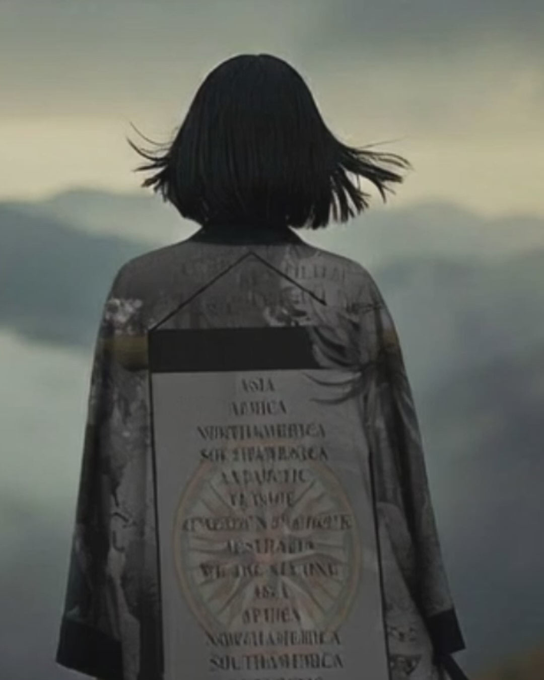 Woman wearing a sophisticated grey luxury kimono with world map print, standing in a cloudy mountain landscape